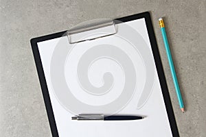 Template of white paper with a ballpoint pen and simple pencil on light grey concrete background in a black tablet with a clip.