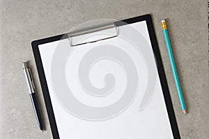 Template of white paper with a ballpoint pen and simple pencil on light grey concrete background in a black tablet with a clip.