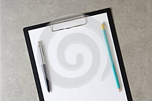 Template of white paper with a ballpoint pen and simple pencil on light grey concrete background in a black tablet with a clip.