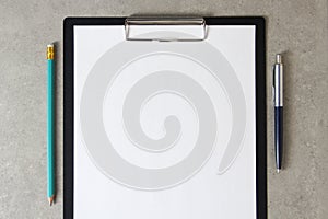 Template of white paper with a ballpoint pen and simple pencil on light grey concrete background in a black tablet with a clip.