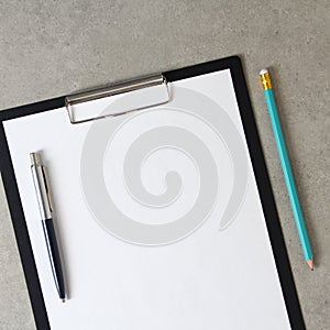 Template of white paper with a ballpoint pen and simple pencil on light grey concrete background in a black tablet with a clip.