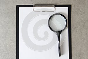 Template of white paper with magnifying glass on light grey concrete background in a black tablet with a clip. Concept