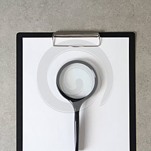 Template of white paper with magnifying glass on light grey concrete background in a black tablet with a clip. Concept