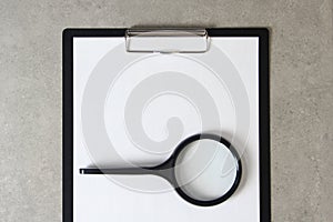 Template of white paper with magnifying glass on light grey concrete background in a black tablet with a clip. Concept of new idea