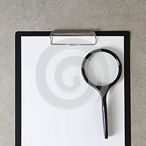 Template of white paper with magnifying glass on light grey concrete background in a black tablet with a clip. Concept of new idea