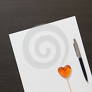 Template of white paper with pen and heart shaped caramel on dark wenge color wooden background