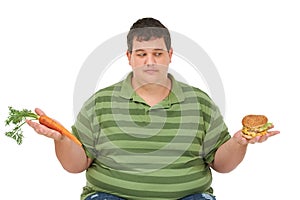 Tempted to have a taste. An obese young man sitting with a carrot in one hand a burger in the other and peering at the