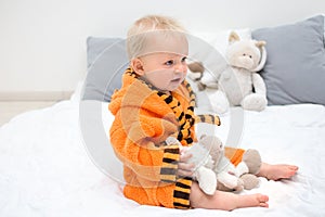 Ten month blond baby boy sitting on bed in orange bathrobe and playing with plush toy. Baby care concept, banner copy space