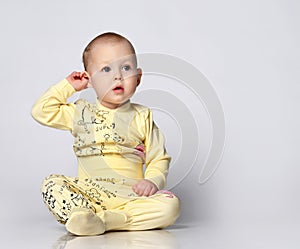 Ten month old baby toddler in a cotton jumpsuit with a print of a cat and funny drawings, sits thoughtfully scratches an
