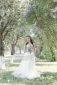 Tender beautiful cute girl bride in a white air dress with a bouquet of lilacs in her hands walking through the park on a sunny sp