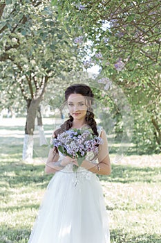 Tender beautiful cute girl bride in a white air dress with a bouquet of lilacs in her hands walking through the park on a sunny sp