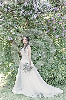 Tender beautiful cute girl bride in a white air dress with a bouquet of lilacs in her hands walking through the park on a sunny sp