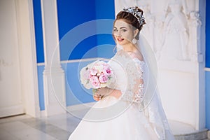 A tender girl with bright curls in a bride`s dress is holding a bouquet of peonies in a blue hall. The bride adorned her