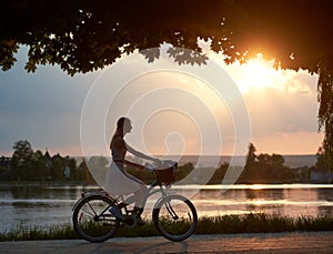 Tender girl rides a retro bike with basket on road near the river on sunset