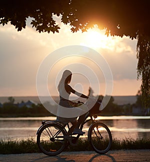 Tender girl rides a retro bike with basket on road near the river on sunset