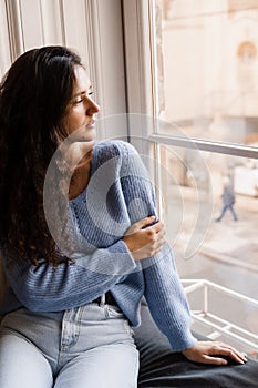 Tender girl is sitting in cafe near big window at cozy winter morning. Cheerful young woman is sitting in cozy