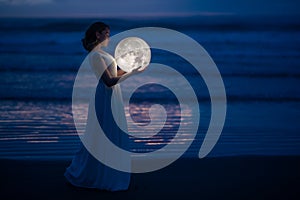 Tender image of a girl. Female magic. Beautiful attractive girl in full growth on a night beach with sand hugs the moon, art photo