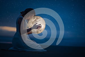 Tender image of a girl; female magic. Beautiful attractive girl on a night beach with sand and stars hugs the moon, art photo. On