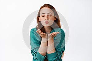 Tender and lovely redhead woman pucker lips, sending air kiss in open hands, standing romantic and cute against white