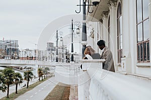 A tender moment as a couple in stylish attire share an embrace on a bridge with a cityscape backdrop and lampposts.