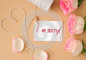 Tender pink roses with spool of white cotton rope in heart shape on beige background. Paper note with text I AM SORRY