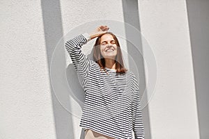 Tender portrait of young caucasian woman with dark hair standing in sunlight outdoors against grey wall, closed eyes gentle smile