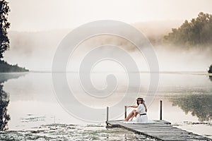 Tender romanitc sentimental female lady in the morning on a wooden pier near the misty river in a white dress
