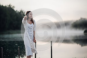 Tender romanitc sentimental female lady in the morning on a wooden pier near the misty river in a white dress