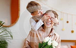 Tender son closes his mother`s eyes, making her a surprise and gives her a bouquet of tulips, congratulating her on mother`s day