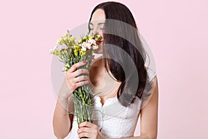 Tender sweet brunette young lady standing straight, holding beautiful flowers in both hands, feeling their spring smell, looks