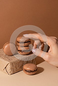 Tender woman fingers touch the brown chocolate macaroons on paper gift box on beige background. Sweet food. Tasty desert. Homemade