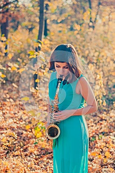 Tender young girl with dark hair in a beautiful long blue dress plays the alto saxophone in a yellow autumn park