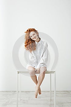Tender young redhead girl in shirt sitting on table over white background smiling looking at camera.