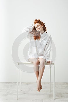 Tender young redhead girl in shirt sitting on table over white background smiling looking at camera correcting hair.