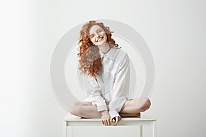 Tender young redhead girl in shirt smiling looking at camera sitting on table posing over white background.