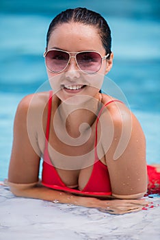 Tenned beautiful asian woman in orange bikini and sunlasses sitting in swimming pool. Fashionable portrait. Elegant