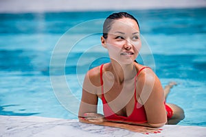 Tenned beautiful asian woman in orange bikini and sunlasses sitting in swimming pool. Fashionable portrait. Elegant