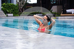 Tenned beautiful asian woman in orange bikini and sunlasses sitting in swimming pool. Fashionable portrait. Elegant