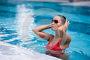 Tenned beautiful asian woman in orange bikini and sunlasses sitting in swimming pool. Fashionable portrait. Elegant