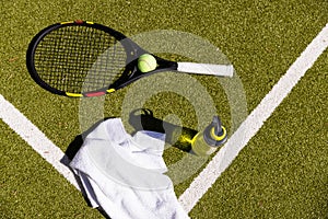 Tennis ball, racket, towel and water bottle lying on sunny outdoor grass tennis court