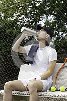 Tennis player drinking water sitting on a bench resting after playing a tennis match on a court in spring time
