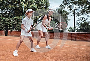 Tennis training. Cheerful mother in sports clothing teaching his daughter to play tennis while both standing on tennis court photo