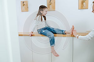 Tense girl sitting on a corridor shelf, her feet being tickled by her mom