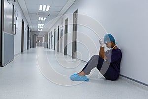 Tensed female surgeon with hands on forehead sitting on floor in the corridor at hospital