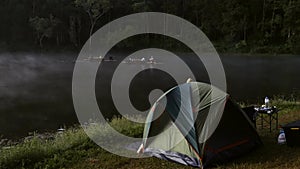 Tent, folding table, and camping gear on fog lake shore in early morning forest