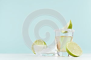 Tequila cocktail with salty rim, piece green lime in shot glass, salt in bowl on soft light pastel blue background.