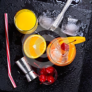 Tequila Sunrise cocktail, orange, ice cubes, maraschino cherries, ice tongs, jigger and straw on a black wet slate tray. Cocktail