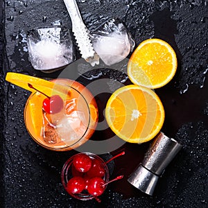 Tequila Sunrise cocktail, orange, ice cubes, maraschino cherries, ice tongs and jigger on a wet black slate tray. Cocktail Set. To