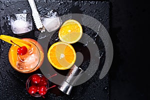 Tequila Sunrise cocktail, orange, ice cubes, maraschino cherries, ice tongs and jigger on a wet black slate tray. Flat lay with co