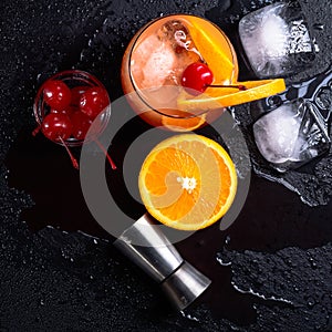 Tequila Sunrise cocktail, orange, ice cubes, maraschino cherries and jigger on a wet black slate tray. Cocktail Set. Top view.